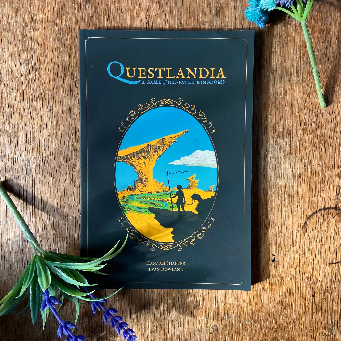 Book titled 'Questlandia' on a wooden surface with decorative faux flowers