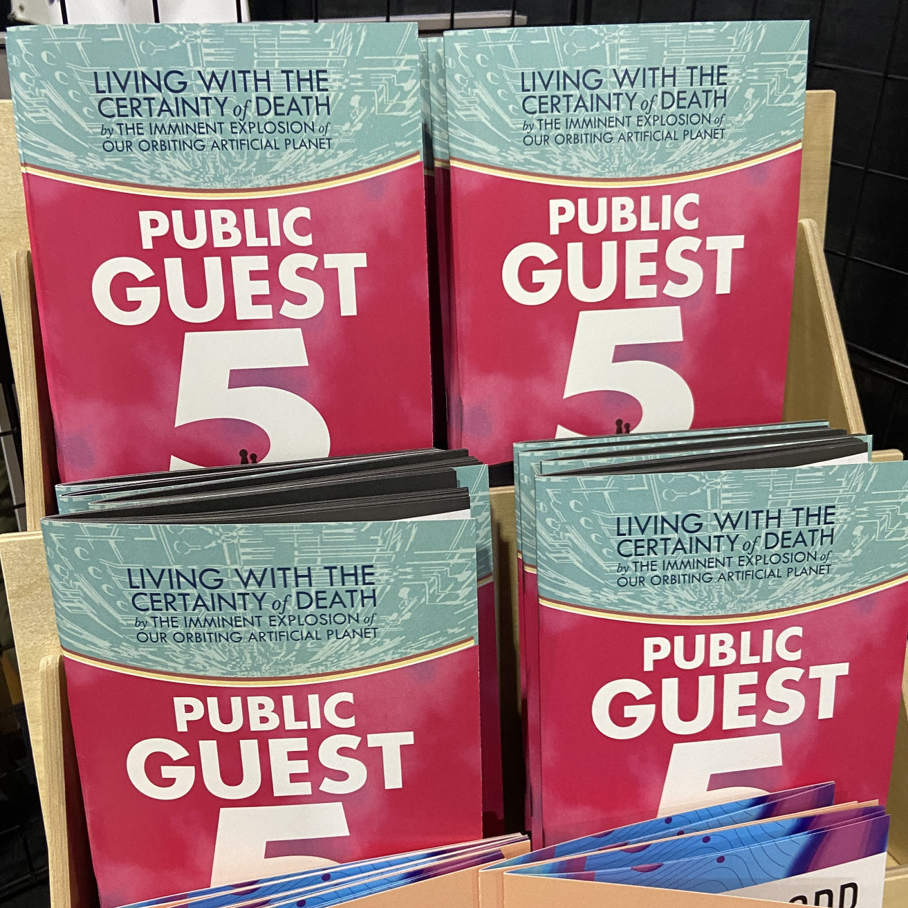 Multiple copies of a book titled 'Public Guest 5: Living with the Certainty of Death by the Imminent Explosion of Our Orbiting Artificial Planet' displayed on a shelf.